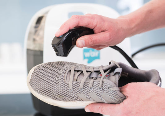 Sneaker Cleaning System to Keep Your Trainers Smelling Clean and Odour Free. A Grey Mesh Trainer is held in a left hand, while the right hand holds the w'air sneaker cleaning device to spray and clean the trainer. 